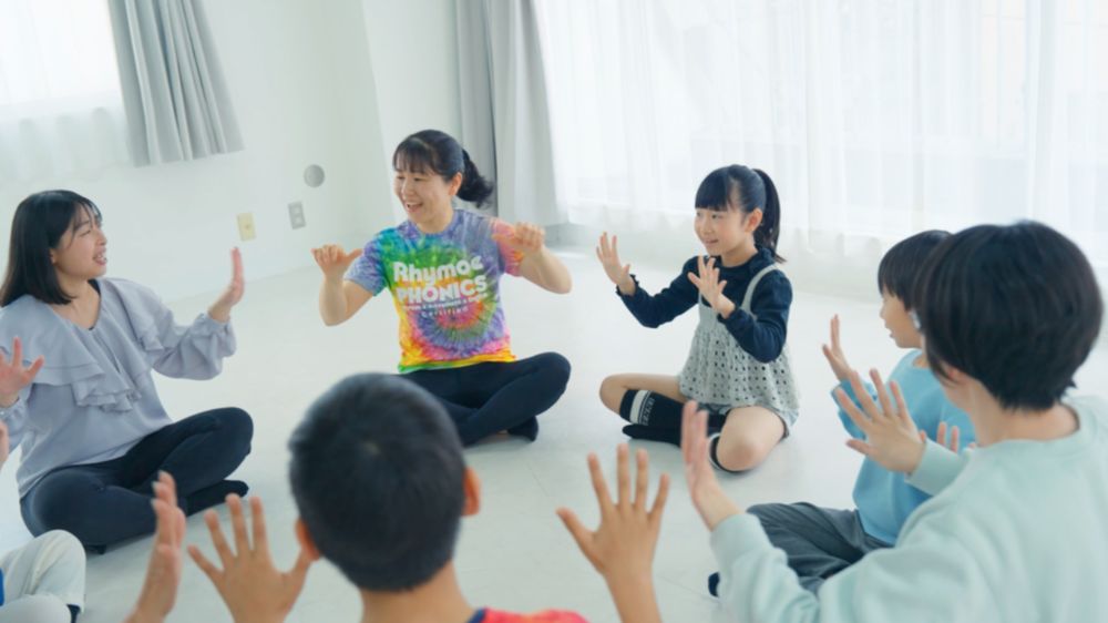 輪になって座り、手を動かしながら英語を声に出す子どもたちと講師